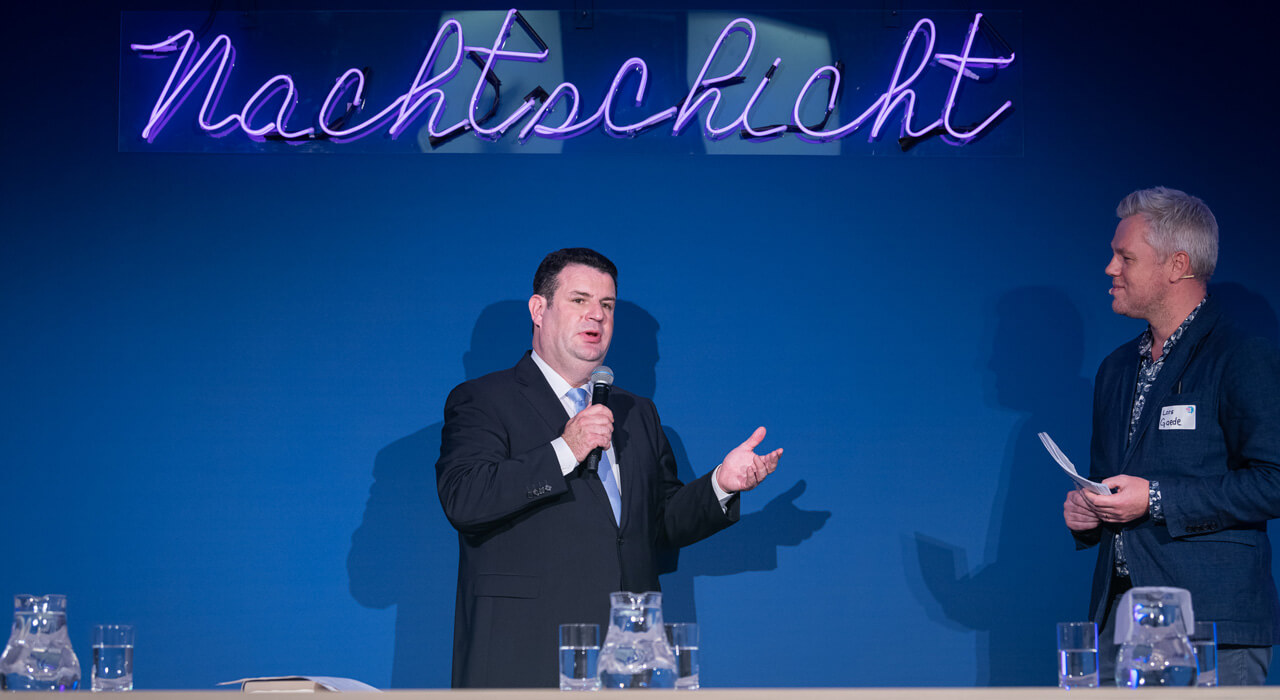 Hubertus Heil spricht in ein Mikrofon und steht neben einem Mann vor einer dunkelblauen Wand mit dem Leuchtschriftzug Nachtschicht.