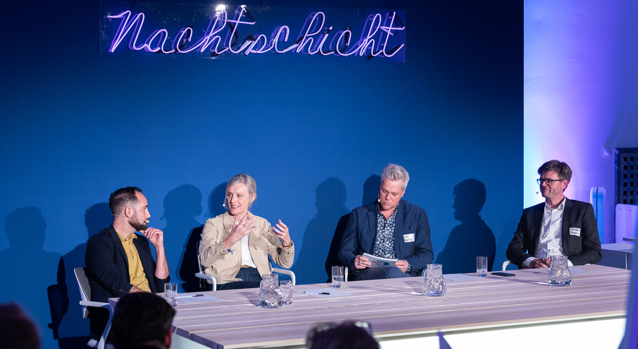 Eine Frau spricht in ein Mikrofon und sitzt mit drei Männern an einem Tisch vor einer dunkelblauen Wand mit dem Leuchtschriftzug Nachtschicht.