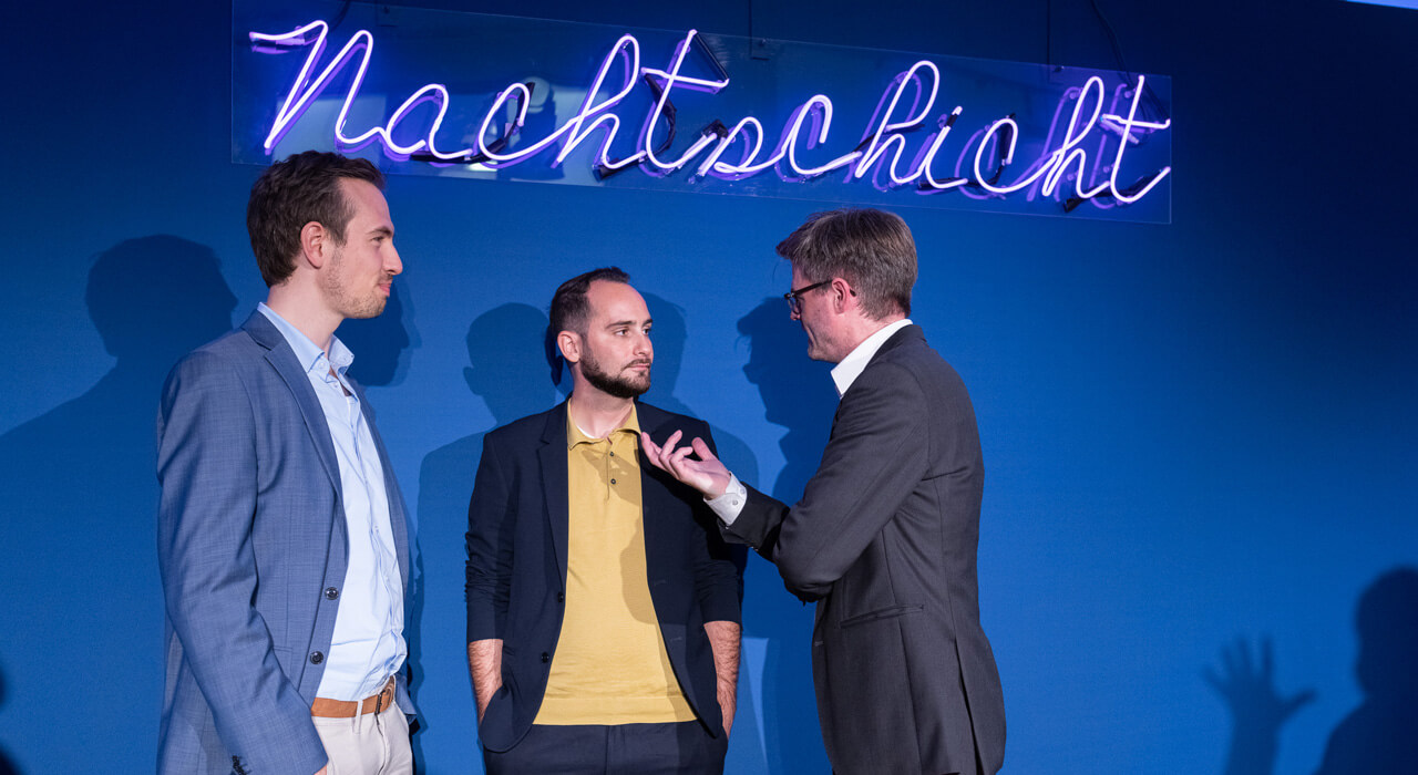 Drei Männer unterhalten sich und stehen vor einer dunkelblauen Wand mit dem Leuchtschriftzug Nachtschicht.