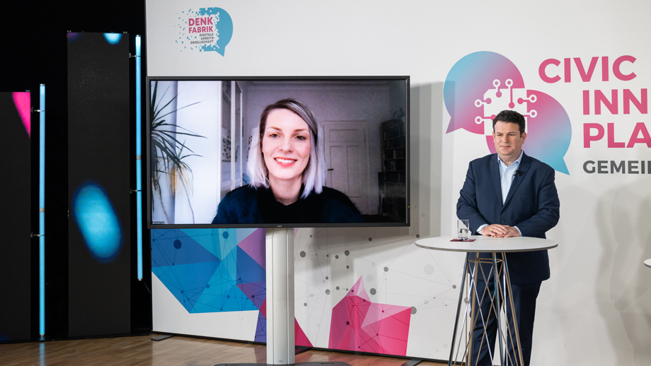 Bundesarbeitsminister Hubertus Heil steht an einem Stehtisch neben einem großen Monitor, auf dem eine Frau zugeschaltet ist.