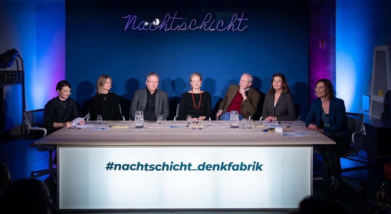 Sieben Personen sitzen an einem Tisch mit der Aufschrift #nachtschicht-denkfabrik vor einer dunkelblauen Rückwand mit dem Schriftzug Nachtschicht.
