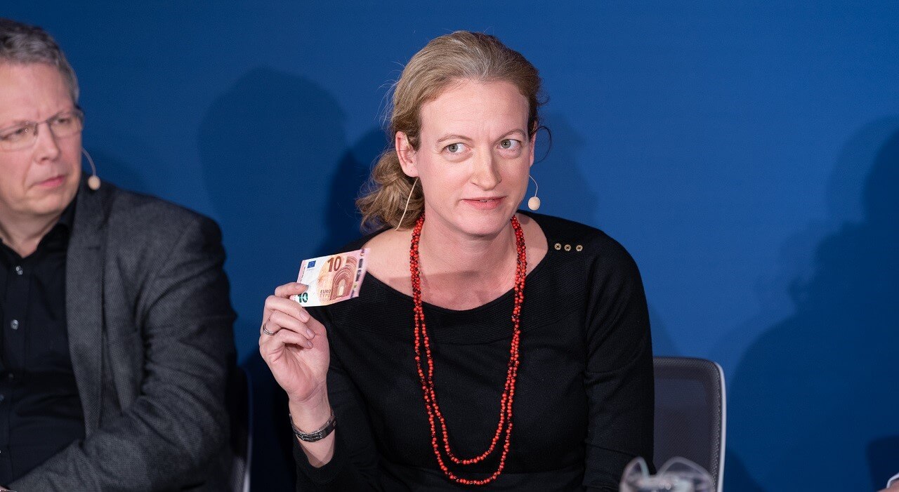 Eine Frau mit Kopfbügelmikrofon sitzt mit weiteren Personen an einem Tisch und hält einen 10-Euro-Schein in der Hand.