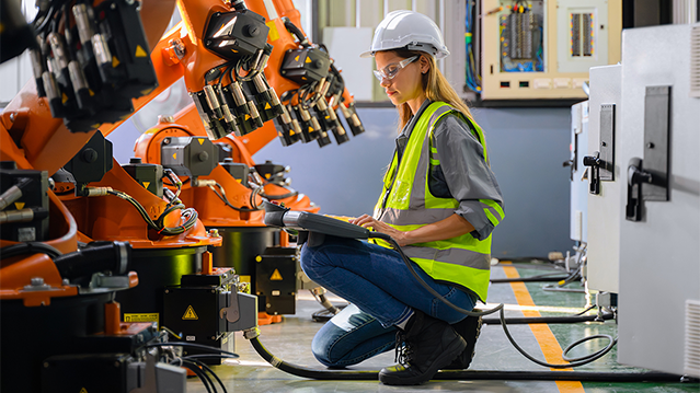 Eine Frau mit Schutzhelm, Schutzbrille und reflektierender Weste bedient ein Bedienfeld, während sie in einer Fabrikanlage neben einem industriellen Roboterarmen kniet.