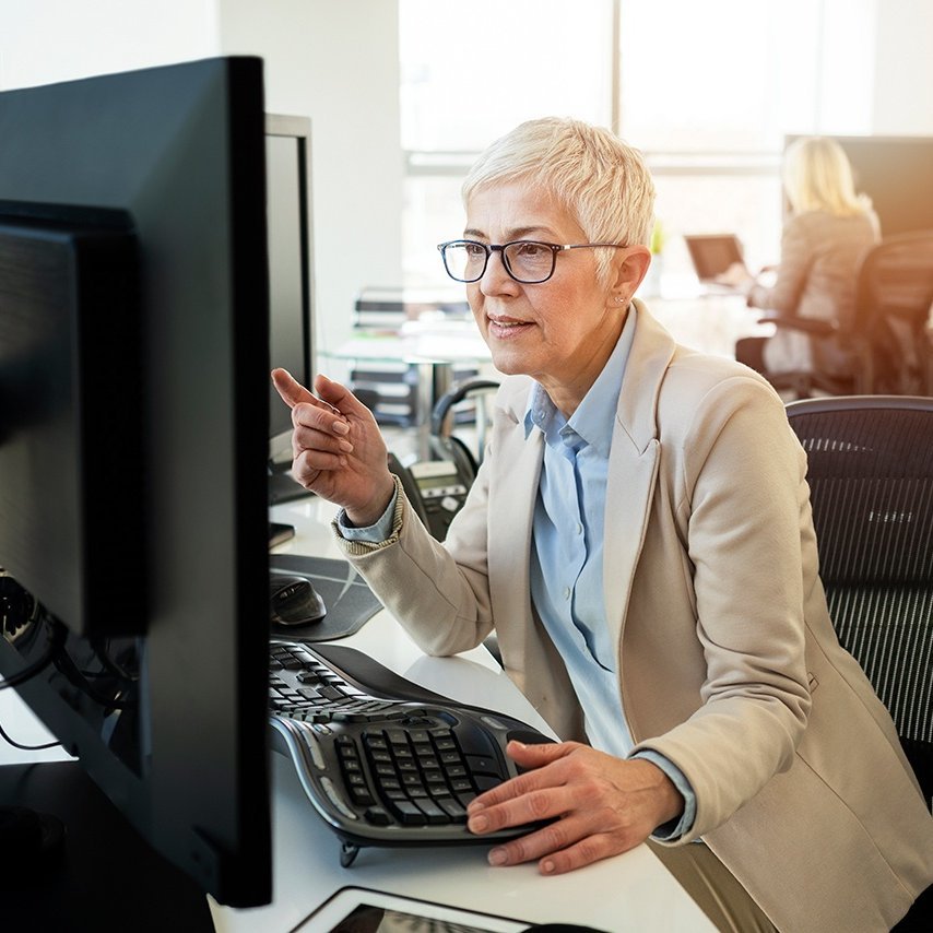 Eine Frau mit Brille sitzt an einem Schreibtisch mit einem Computer.