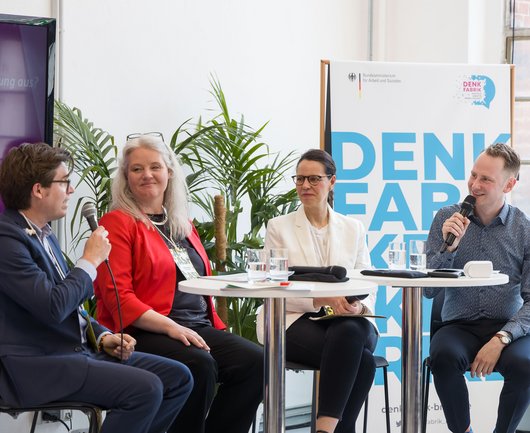 Jakob Kort, stellvertretender Leiter der Abteilung Digitalisierung und Unternehmensentwicklung, spricht mit Ana Dujíc, Abteilungsleiterin der Denkfabrik, Professorin Katharina Zweig und Moderator Mads Pankow. Im Hintergrund sind ein Plakat der Denkfabrik und Pflanzen.