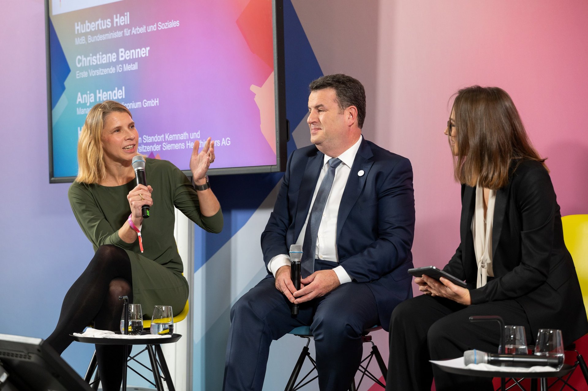 Anja Hendel, hält ein Mikrofon in der Hand und spricht. Rechts neben ihr sitzen Hubertus Heil und die Moderatorin der Diskussion, Larissa Holzki.