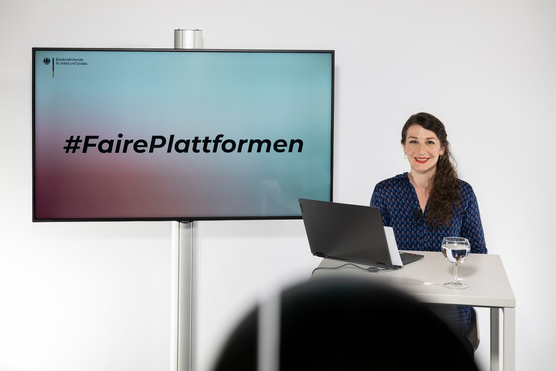 Eine Frau steht mit einem Laptop an einem Stehtisch vor einem Monitor mit dem Titel #FairePlattformen.